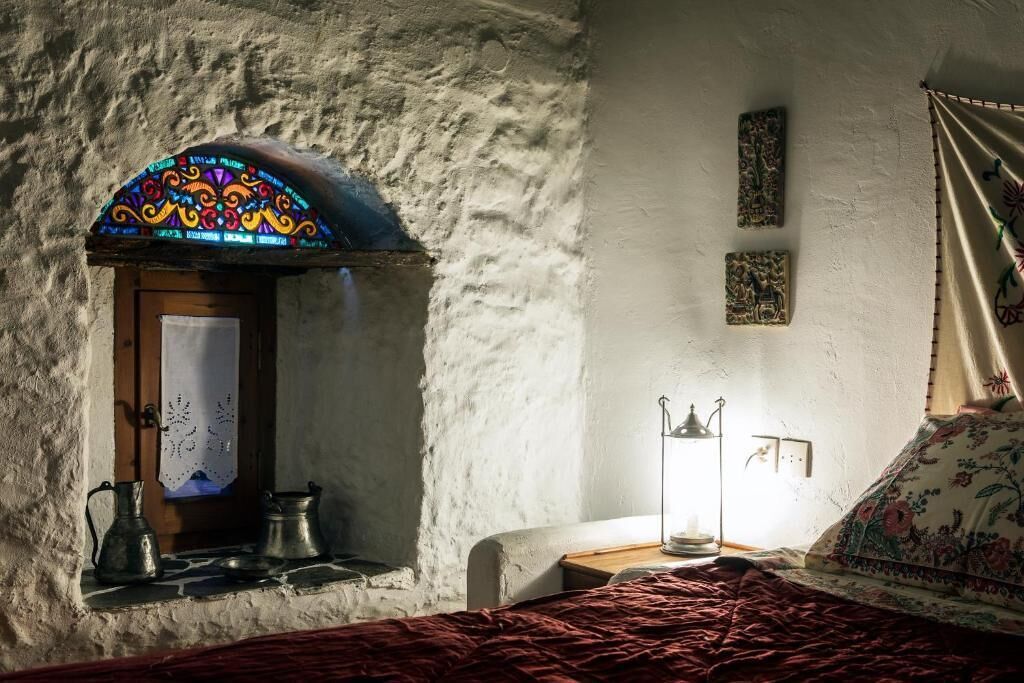 Une pièce rustique avec un mur en pierre, une petite fenêtre ornée d’un vitrail aux couleurs vives, un lit avec un oreiller à motifs floraux et une table de chevet avec une lampe et deux récipients en métal.
