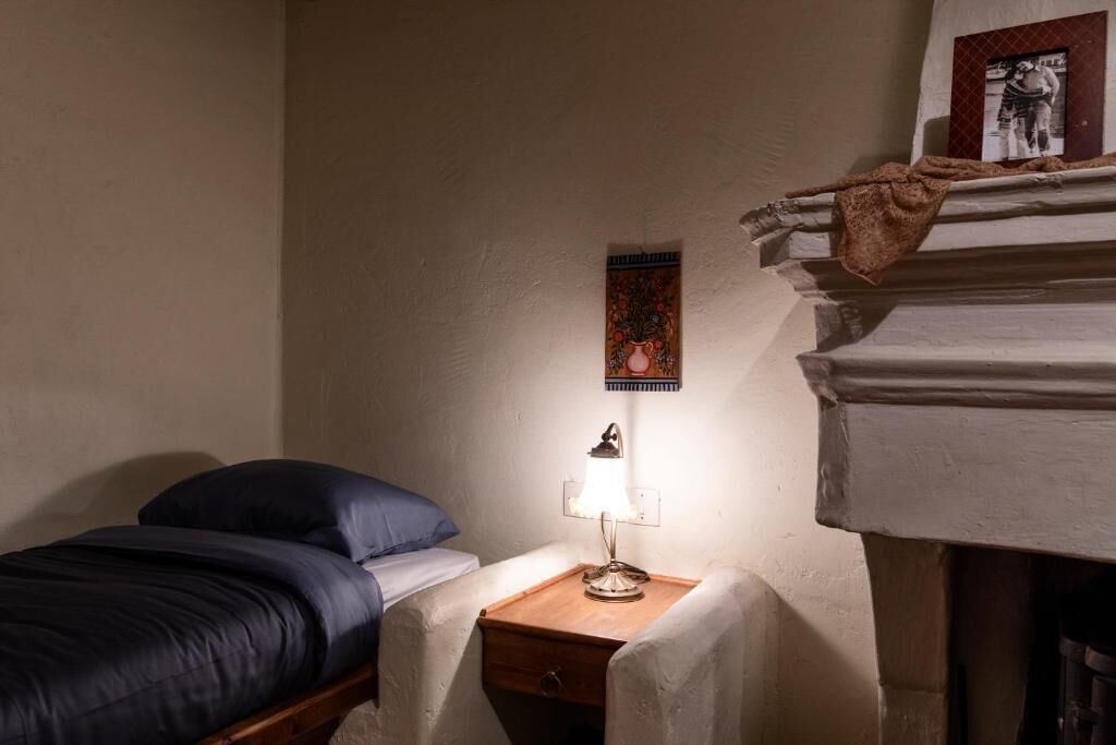 Ein kleiner, gemütlicher Raum mit einem Bett, einem Nachttisch mit Lampe und einem gerahmten Bild an der Wand.