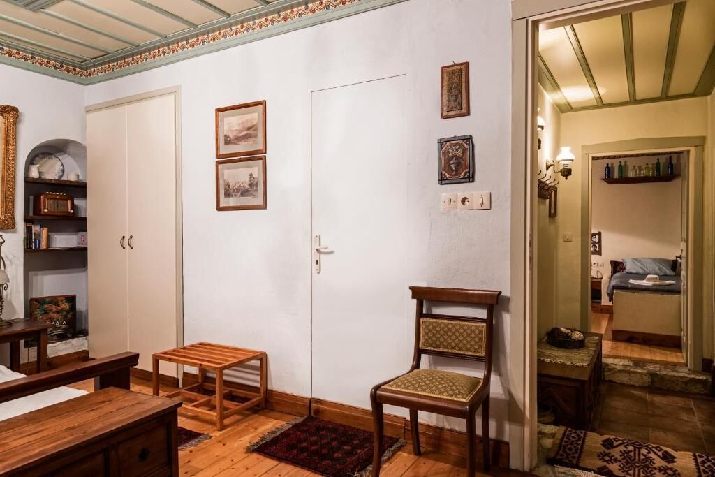 Ein Zimmer mit Holzmöbeln, einem Sessel, einem kleinen Tisch und einer Tür. An der Wand hängen Bilder in Rahmen und ein Regal mit Büchern und Dekorationsobjekten.