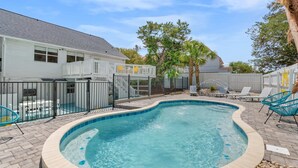 Une piscine à bord incurvé, entourée d’une clôture blanche, et un espace patio aménagé avec des chaises longues et une petite table.