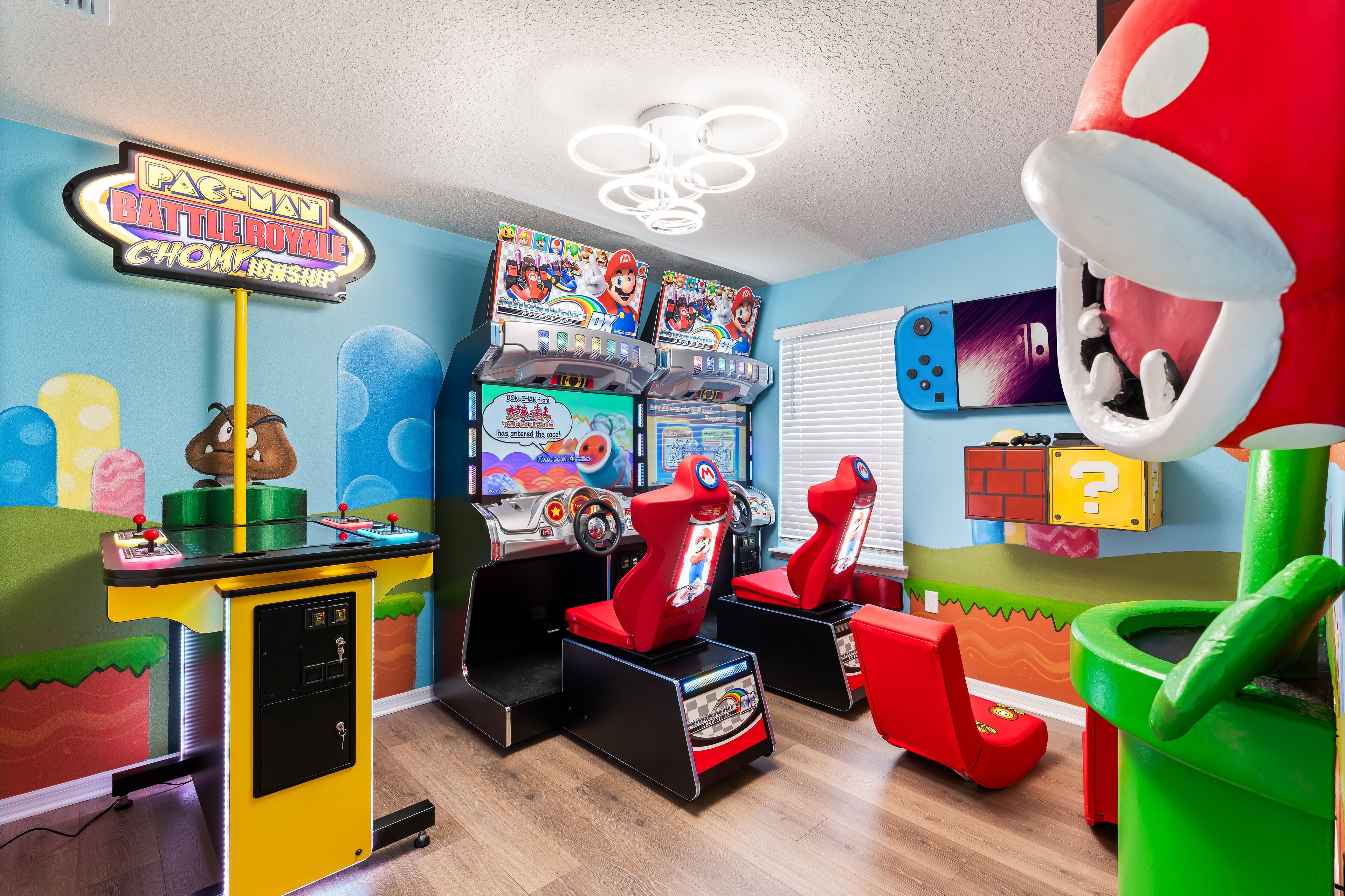 A brightly colored arcade room with Pac-Man and Mario game machines, a neon sign, and a large plush toy.