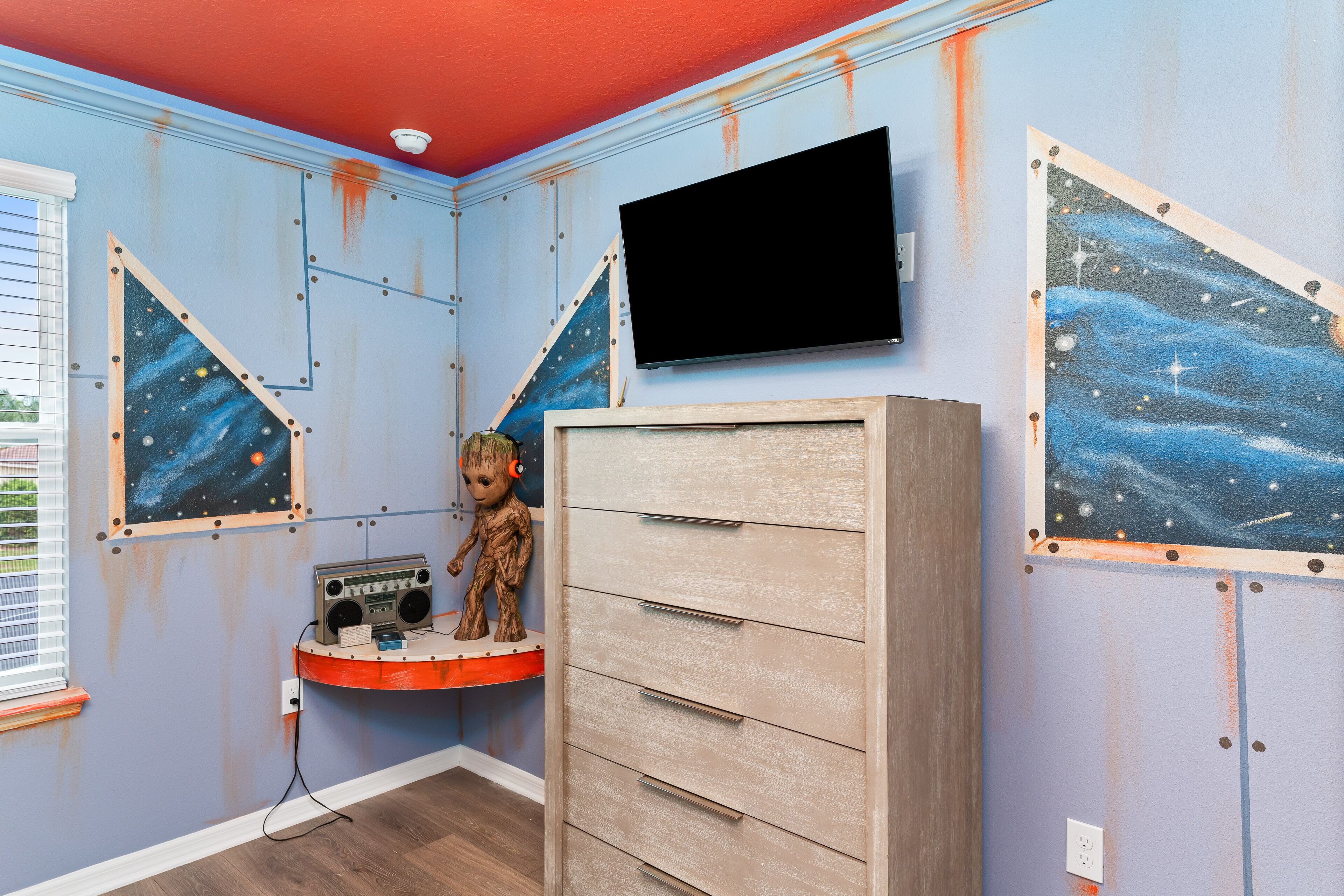 Una habitación con un televisor montado en la pared, un armario de madera y un estante con una radio y un juguete.