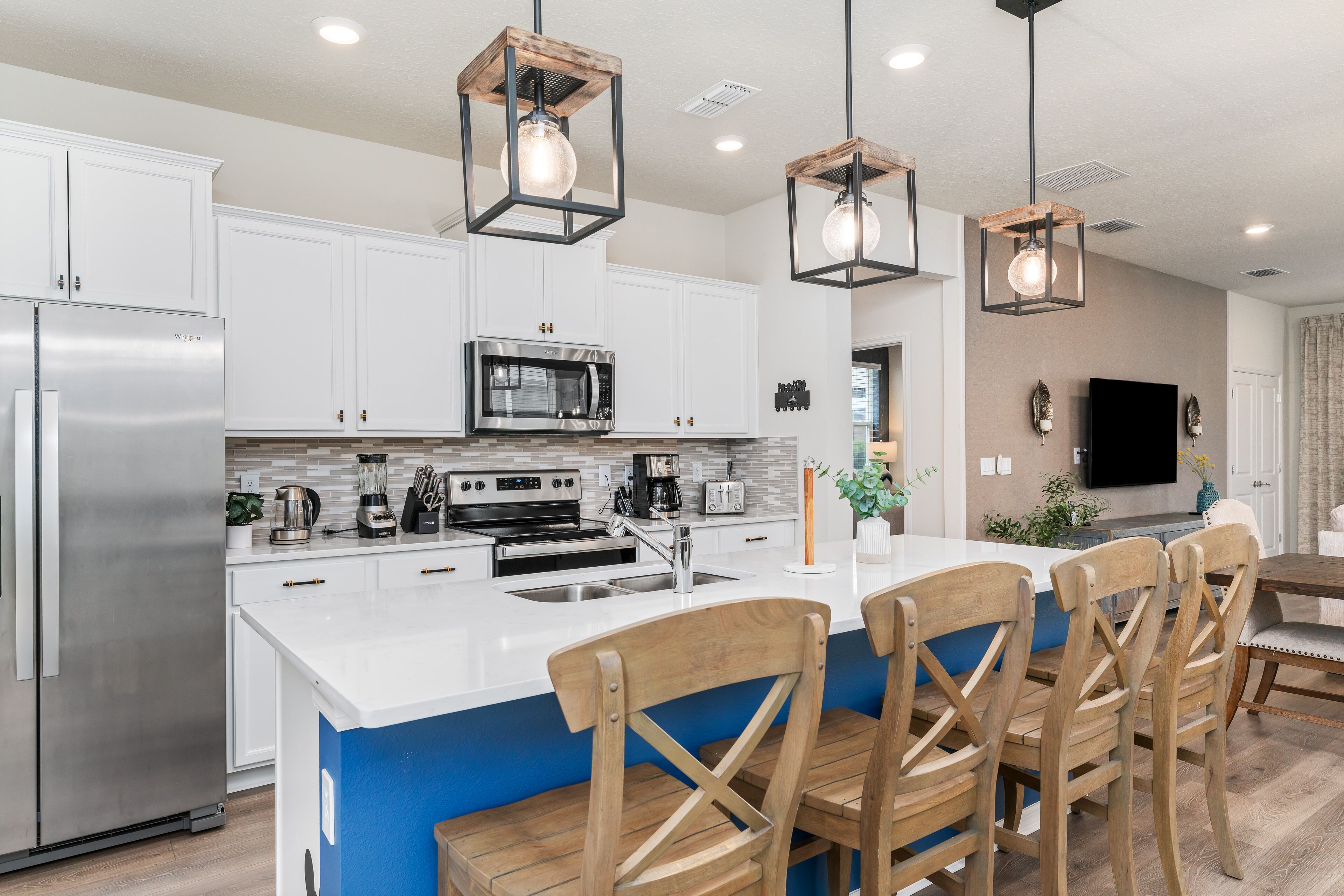 Una cocina moderna con armarios blancos, electrodomésticos de acero inoxidable, una isla central con taburetes y luminarias colgantes.