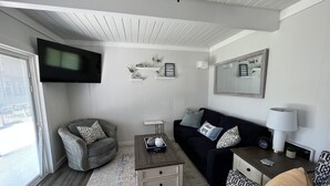 Sala de estar com poltrona cinza, sofá preto, mesa de centro e um banquinho estampado. Há uma TV de tela plana na parede, uma mesinha com abajur e uma janela com persianas.