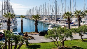 A marina with numerous boats docked along the water, a parking area with cars, and palm trees lining the shore.