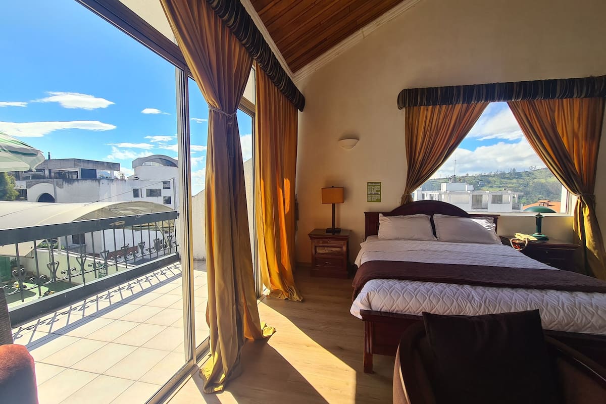 Habitación de hotel con balcón, cama, mesita de noche y vista panorámica de la ciudad.