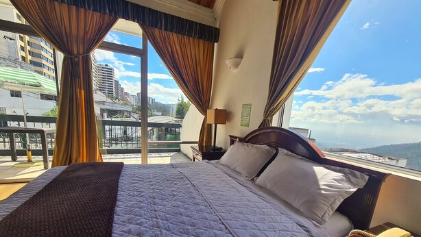 A hotel room with a large bed, a bedside telephone, and a view of the cityscape through large windows. - Hotel & Suites Gonsalez Suarez Quito (Quito)