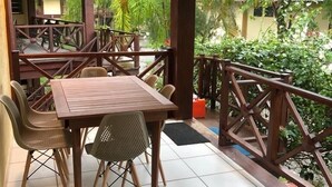 Un espacio comedor al aire libre con techo, que incluye una mesa y sillas de madera, rodeado por barandilla de madera y abundante vegetación.