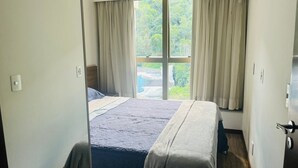 A hotel room with a bed, curtains, and a window with a view of greenery. - Flat near the river Center/Barra Olímpica (Rio de Janeiro)