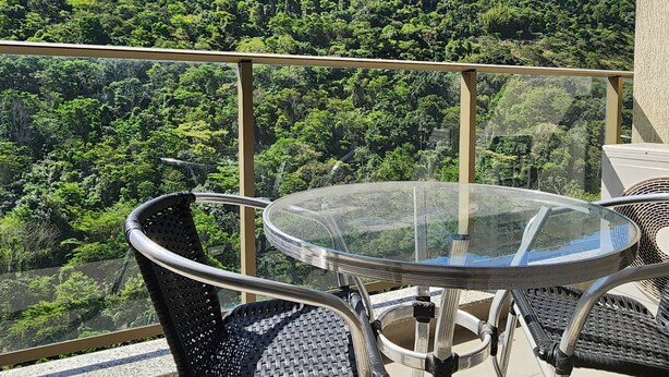 A balcony with a glass table and chairs, overlooking a lush green forested hillside. - Flat near the river Center/Barra Olímpica (Rio de Janeiro)