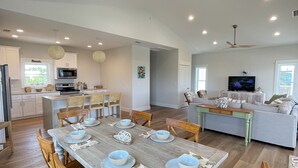A spacious dining area with a wooden table set for six, surrounded by wooden chairs. A kitchen with white cabinets and a microwave is visible in the background. - The Beach Lily - South Cape San Blas - Pet Friendly (Port St. Joe)