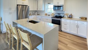 A modern kitchen with white cabinets, a stainless steel refrigerator, a large island with bar stools, and pendant lighting. - The Beach Lily - South Cape San Blas - Pet Friendly (Port St. Joe)
