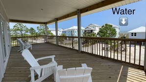 A covered wooden deck with white Adirondack chairs, overlooking a beach and houses. - The Beach Lily - South Cape San Blas - Pet Friendly (Port St. Joe)