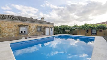 Una piscina con paredes de piedra y techo de tejas, rodeada de un jardín y un edificio con puerta y ventana.