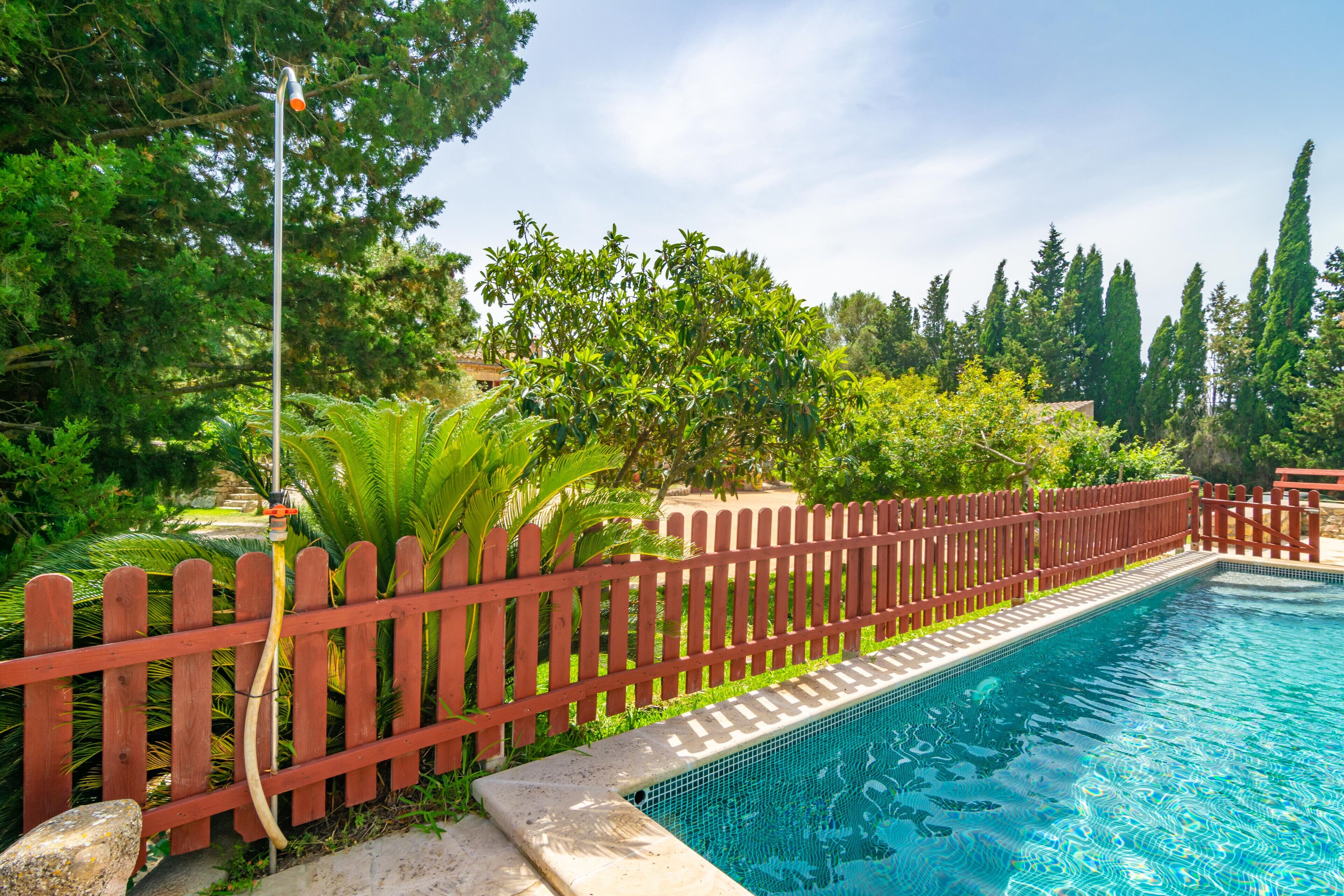 Una piscina con valla de madera roja, rodeada de abundante vegetación y altos árboles.