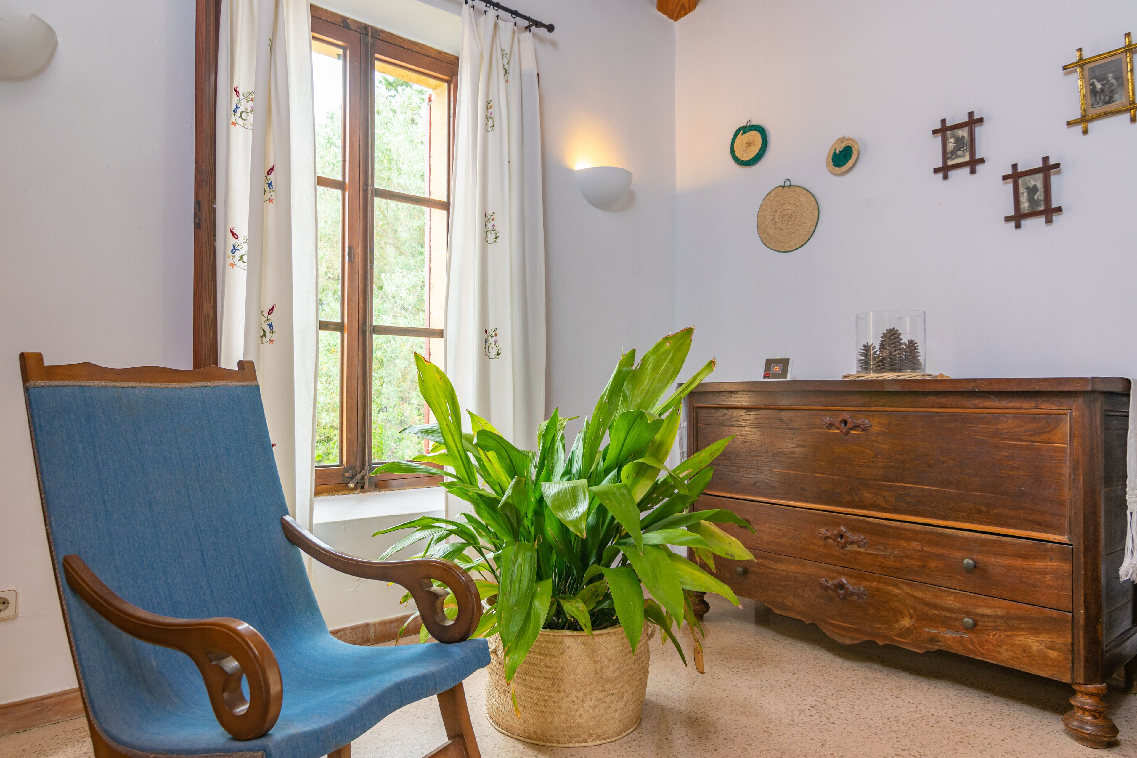 Una habitación con una silla de madera, una planta en maceta, un aparador de madera y una ventana con cortinas.