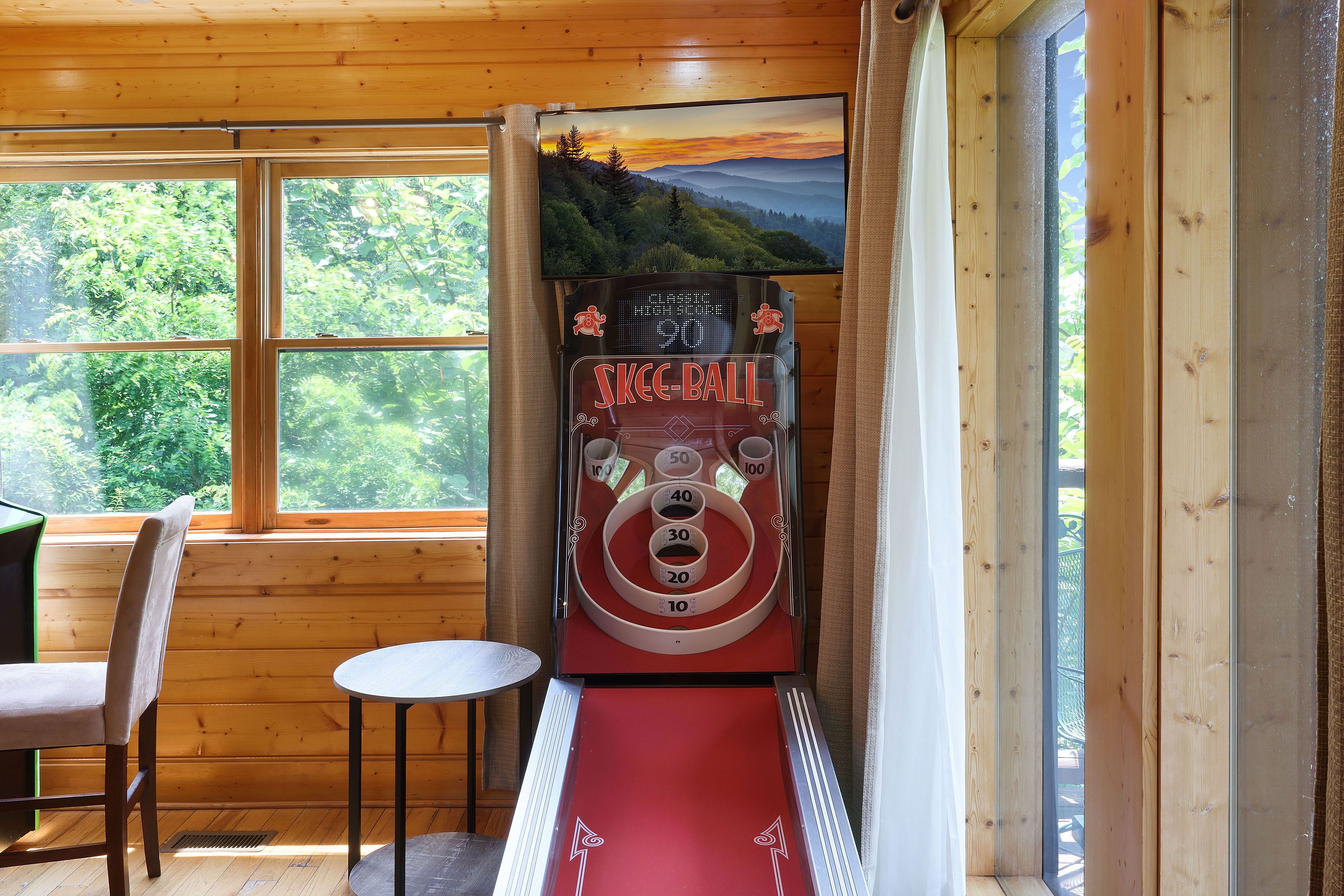 A room with a pinball machine, a chair, and a window with a view of greenery.