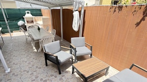 Un patio al aire libre con una mesa blanca, dos sillas grises acolchadas y un banco de madera.