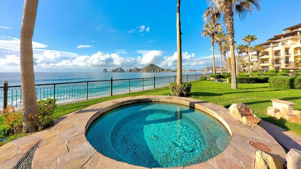 Une piscine à l’eau d’un bleu limpide, entourée de dalles de pierre et de palmiers, qui offre une vue sur une plage et l’océan, avec une clôture visible au loin.