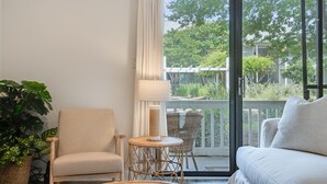 Een kamer met een bank, een stoel, een klein rond tafeltje, een lamp en een groot raam met uitzicht op groen en gebouwen.