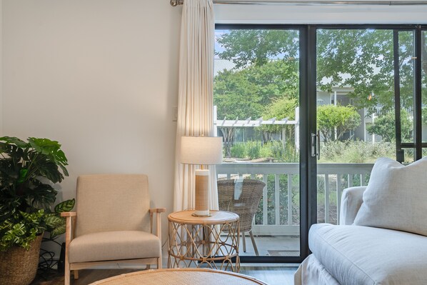 A room with a sofa, a chair, a small round table, a lamp, and a large window with a view of greenery and buildings.
