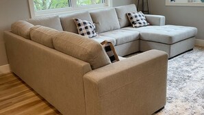 A beige sectional sofa with checkered pillows, positioned in a room with a large window and a wooden floor. - Entire Top Floor Only: Okoboji Oasis/ Private Entrance (Arnolds Park)