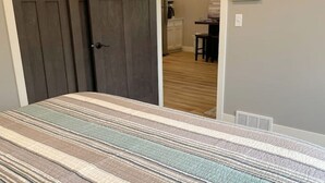 A bedroom with a striped bedspread, a closed door, and a kitchen area visible through an open door. - Entire Top Floor Only: Okoboji Oasis/ Private Entrance (Arnolds Park)