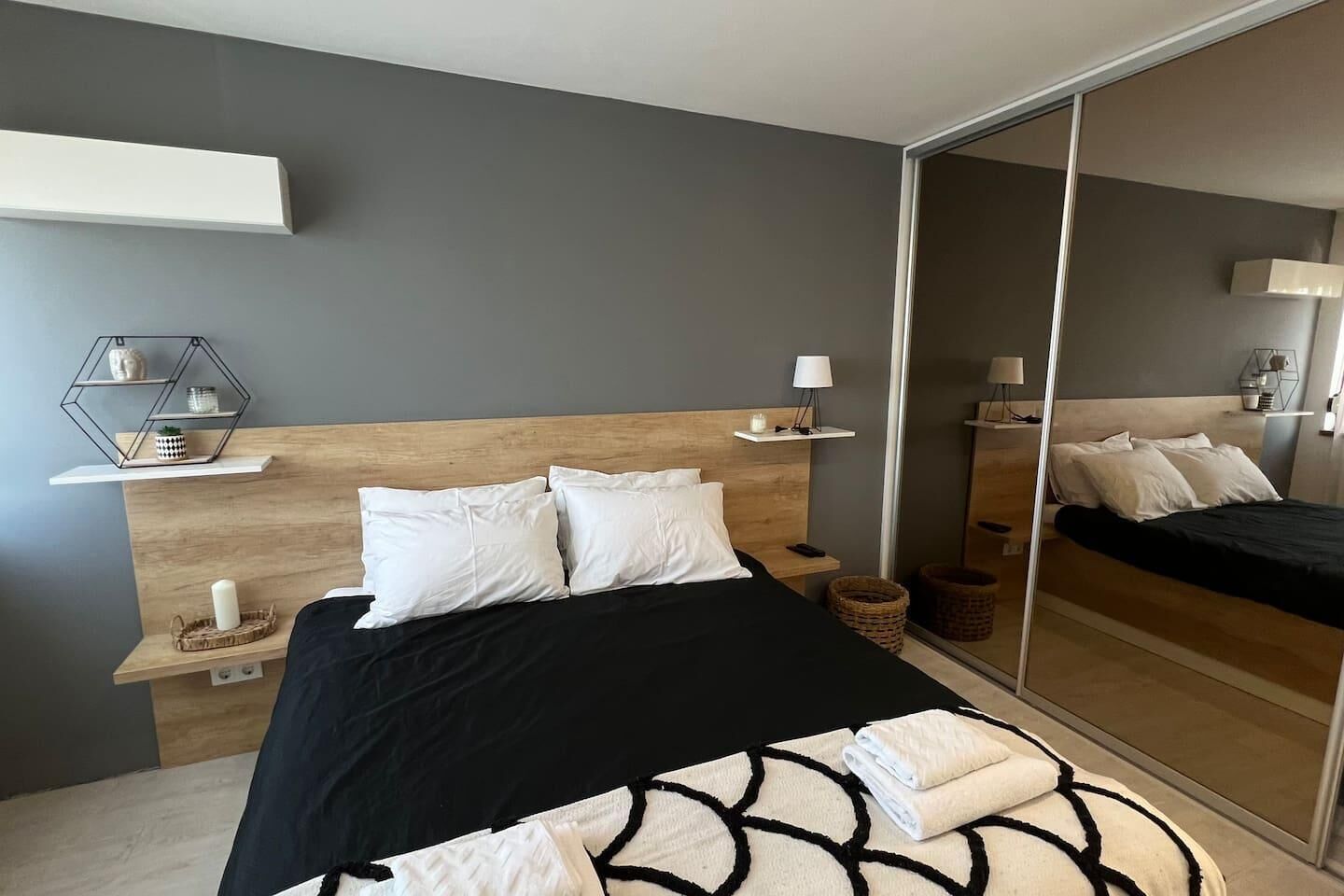 A modern bedroom with a wooden headboard, a black and white patterned bedspread, and a large mirror reflecting the room.