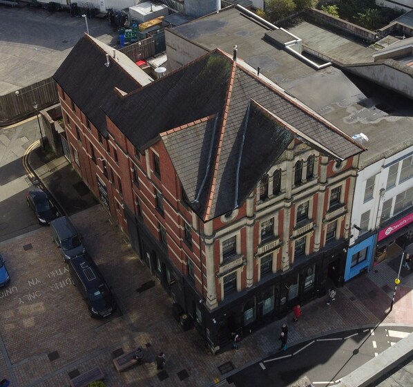 A multi-story building with a prominent corner entrance, a visible ground floor with a shop, and a parking area with cars. - Captivating 1-Bed apartment, Merthyr Tydfil - (Merthyr Tydfil)