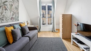 A modern loft apartment with a gray sofa, a large window with curtains, a flat-screen TV, and a wooden cabinet. - Lovely studio for 2 people in Paris (Paris)