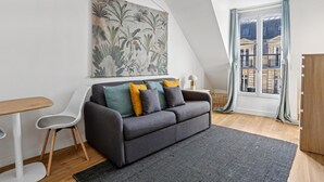 A modern loft apartment with a grey sofa, a white chair, a small table, and a large window with a view of buildings. - Lovely studio for 2 people in Paris (Paris)