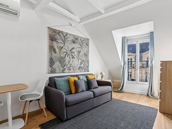A modern loft apartment with a grey sofa, a white chair, a small table, and a large window with a view of buildings. - Lovely studio for 2 people in Paris (Paris)