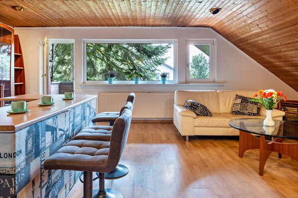 A loft-style room with a wooden ceiling, a bar area with stools, a sofa, and a coffee table with a vase of flowers.
