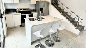 A modern kitchen with white cabinetry, stainless steel appliances, a central island with bar stools, and a staircase with a metal railing.