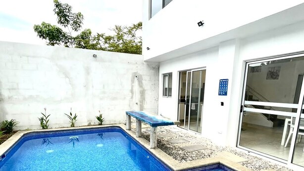 A rectangular swimming pool with blue mosaic tiles, surrounded by a concrete deck and a white wall.