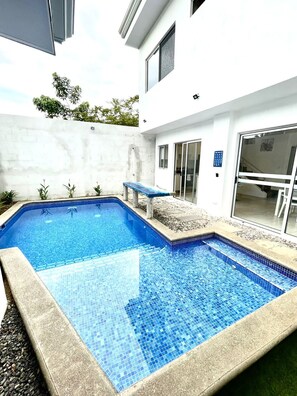 Una piscina rectangular con mosaicos azules, rodeada por un solárium de concreto y una pared blanca.