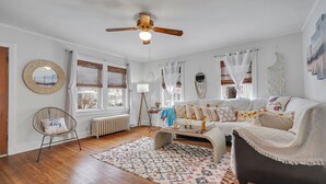 A cozy living room with a white sofa, a wooden floor, a ceiling fan, and a large window with curtains. - Gorgeous home walking distance to the beach (Rochester)