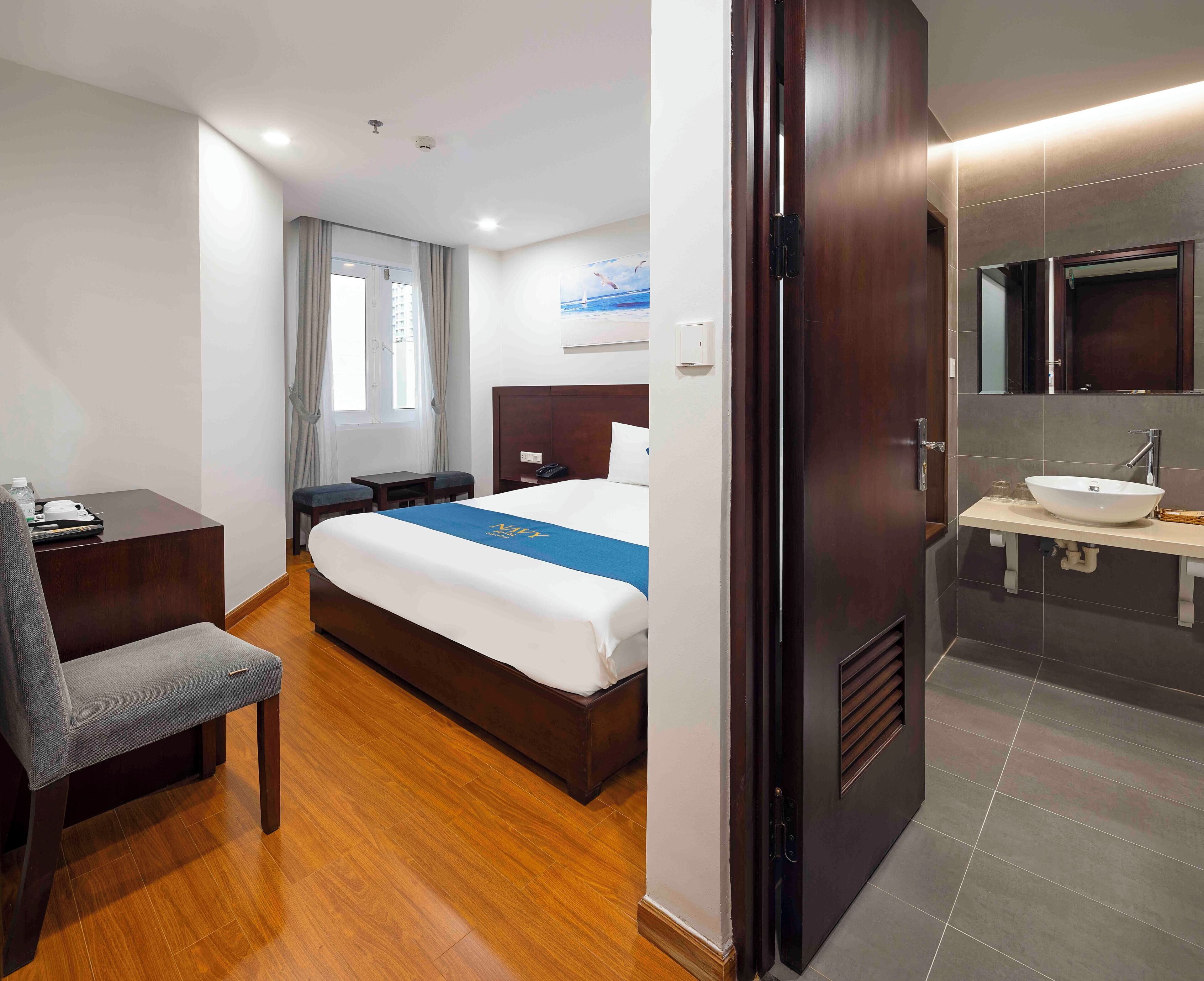 A hotel room with a bed, a desk, a chair, a bathroom with a sink and mirror, and a window with curtains.