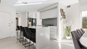 A modern kitchen with white cabinetry, a central island with bar stools, and a dining area with a table and chairs. - Modern Port Coogee Luxury Home (North Coogee)