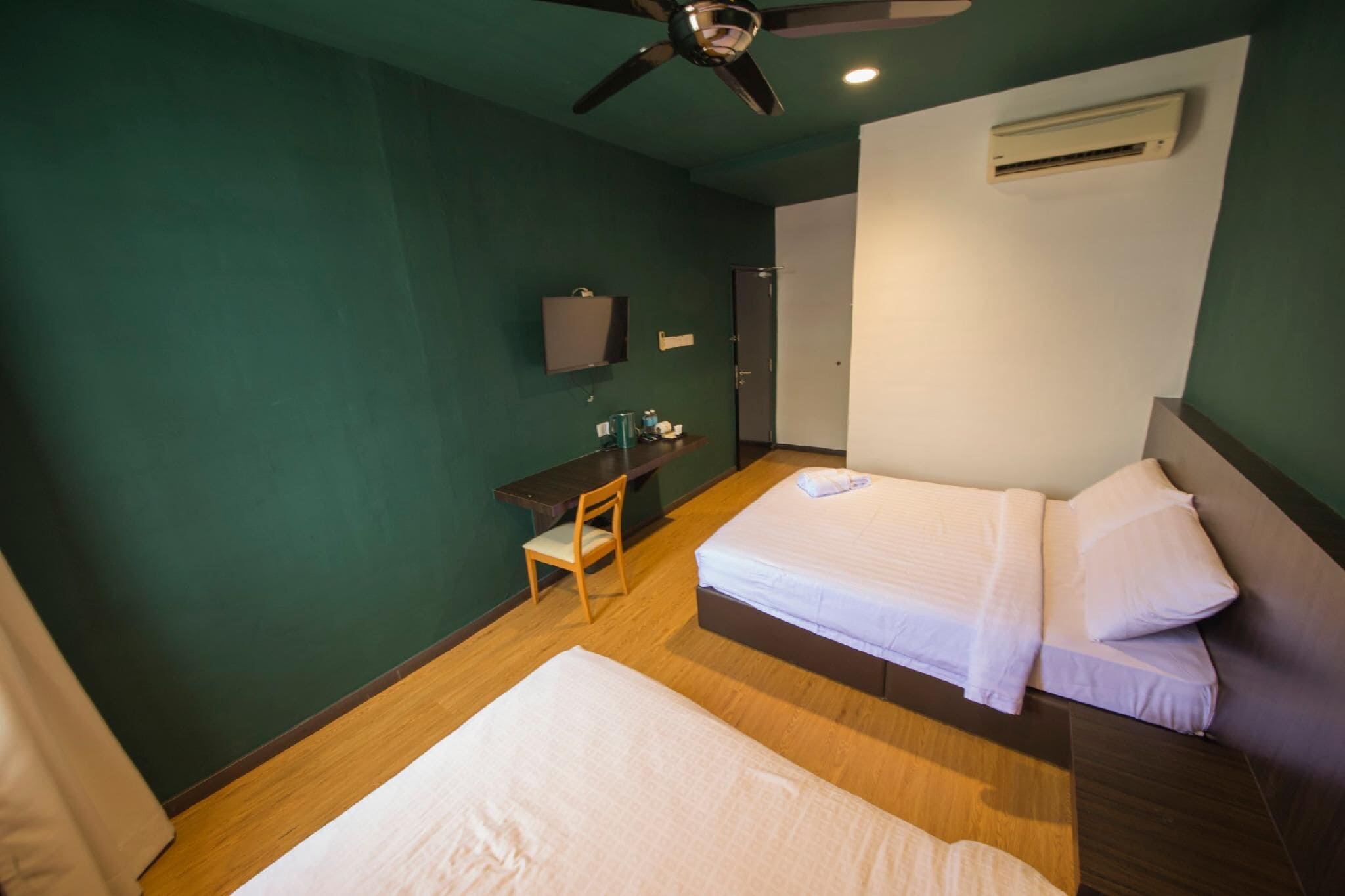 A hotel room with two beds, a desk, a chair, a ceiling fan, and an air conditioning unit.