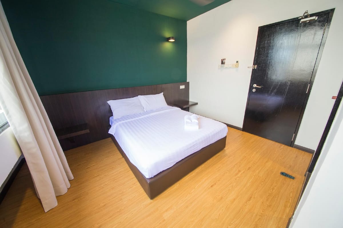 A hotel room with a bed, wooden floor, green walls, a black door, and a small bedside table.