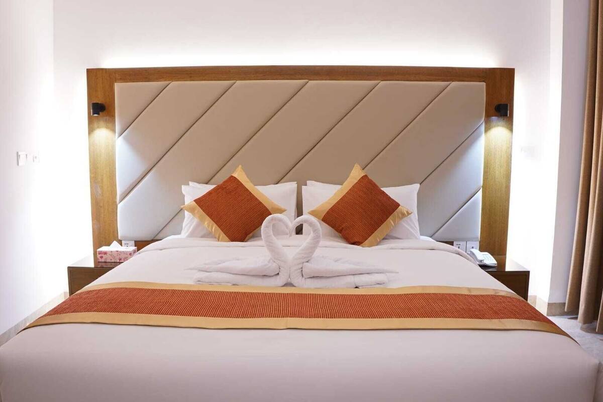 A hotel room with a bed featuring a heart-shaped towel arrangement, two pillows, and a wooden headboard.