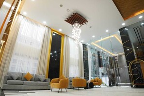 A modern hotel lobby with a grey sofa, two mustard-colored armchairs, a chandelier, and a glass wall with a view of the city. - Studio23 (Dhaka)