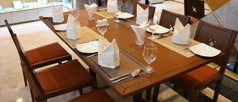 A dining table set with white napkins, silverware, and wine glasses, surrounded by wooden chairs.