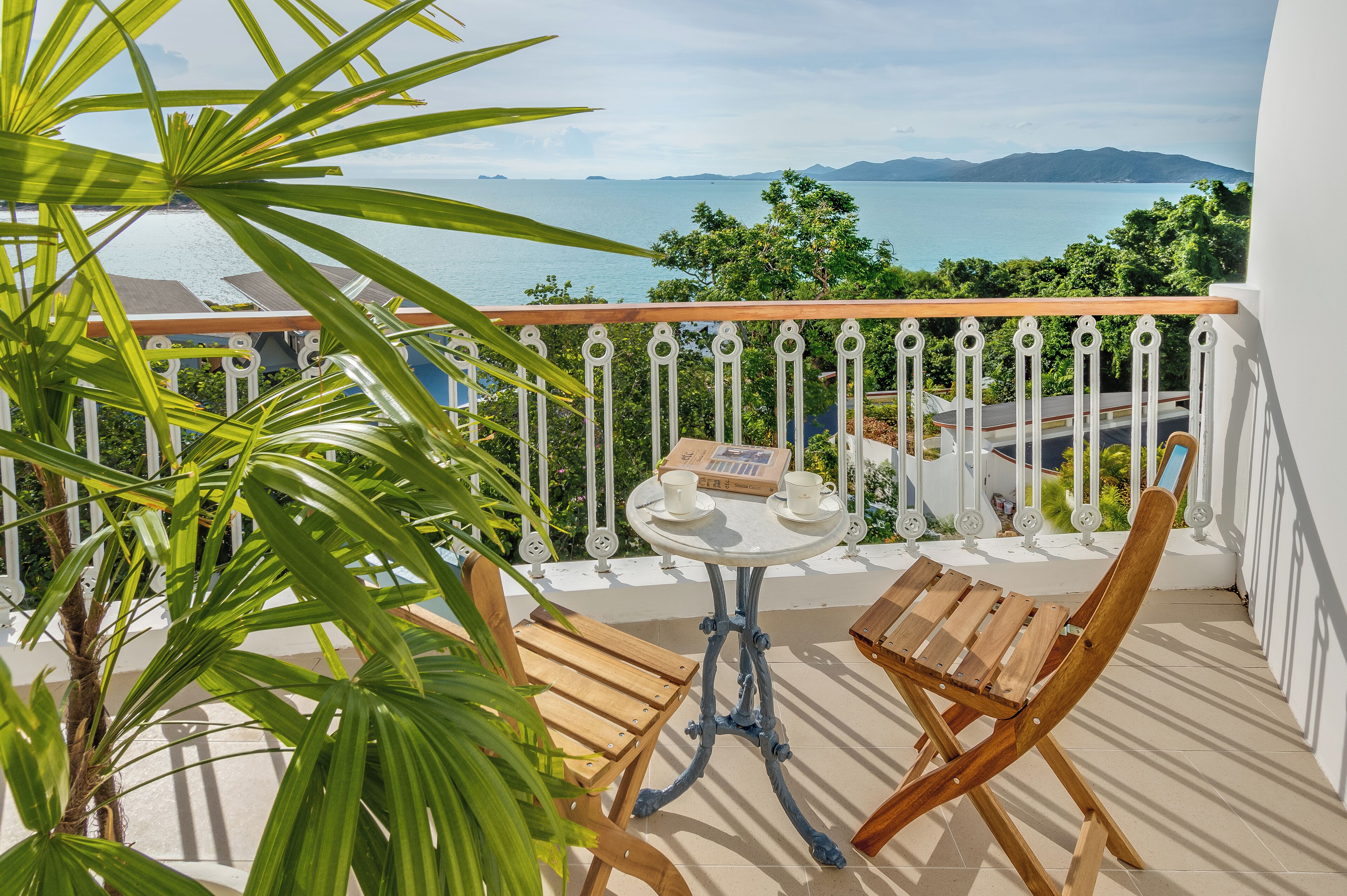 A balcony with a wooden chair, a small table with two cups, and a view of the sea and mountains.