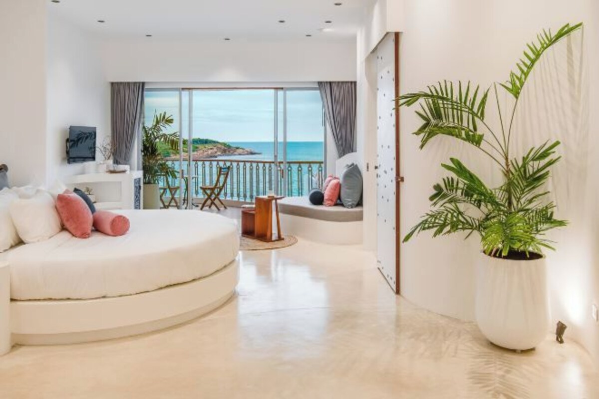 A modern living room with a curved white sofa, a coffee table, and a large potted plant. A balcony with a view of the sea is visible through the large windows.
