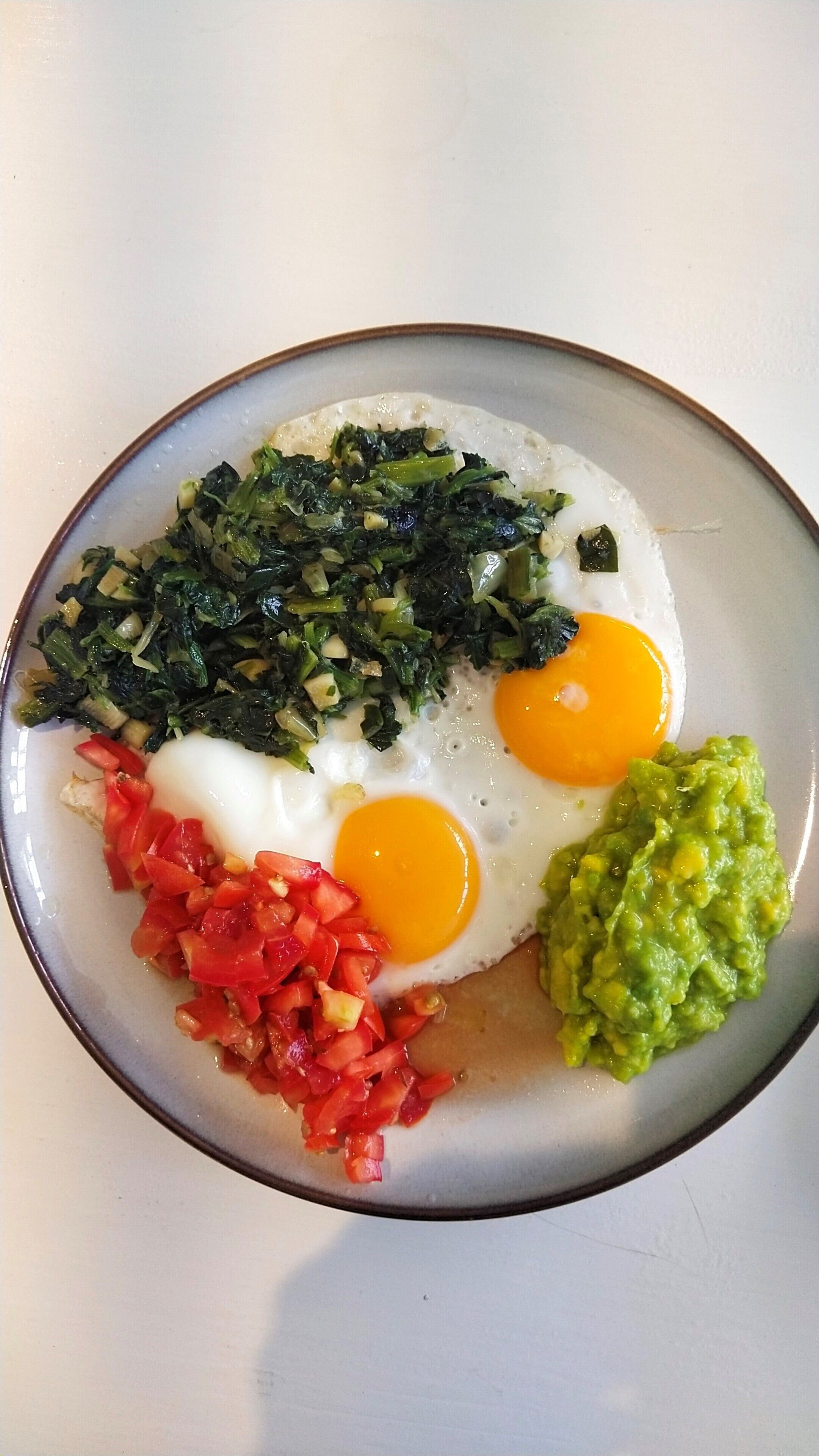 A plate with two sunny-side-up eggs, sautéed greens, diced tomatoes, and guacamole.