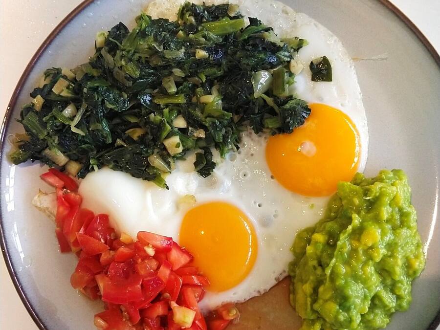 A plate with two sunny-side-up eggs, sautéed greens, diced tomatoes, and guacamole.