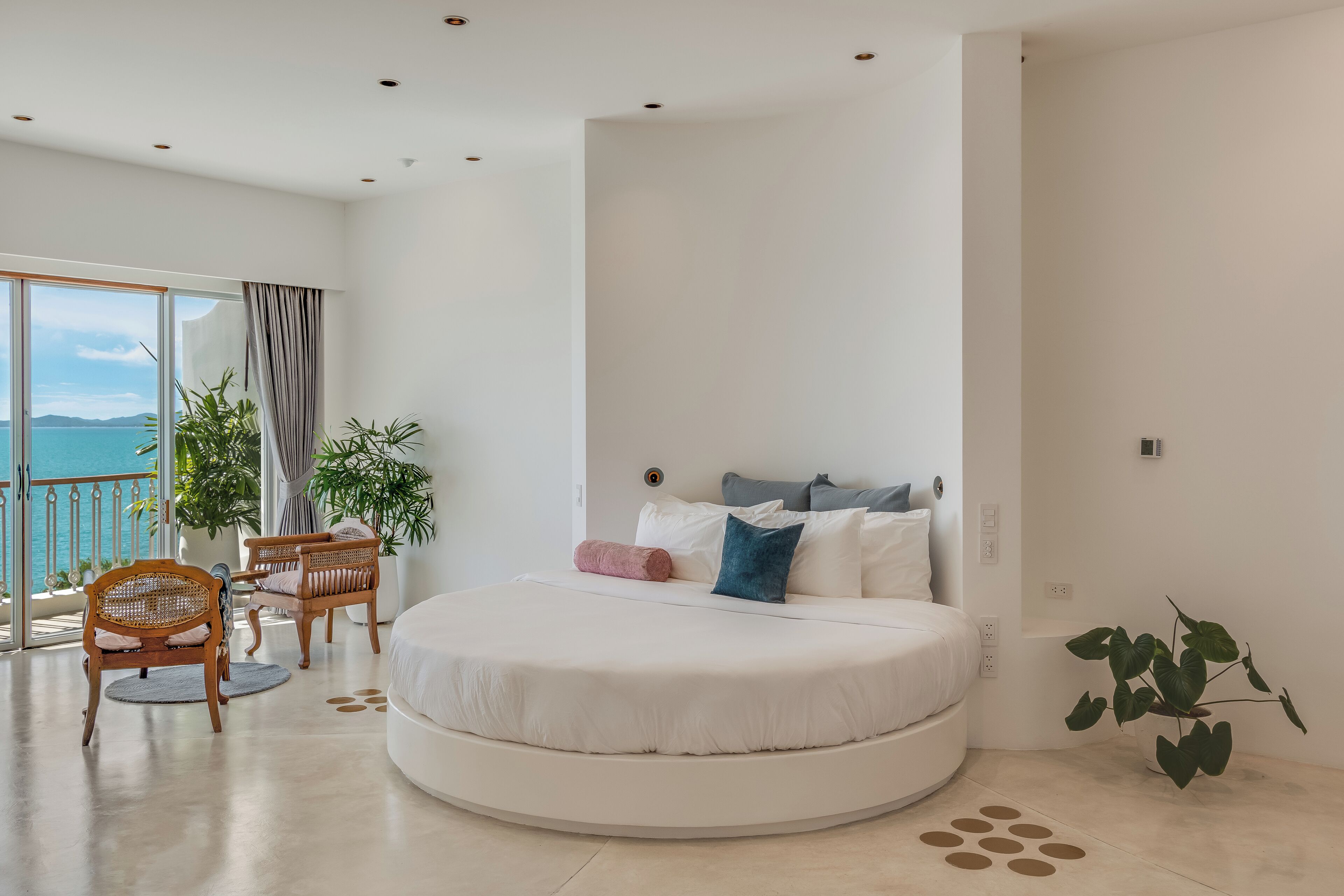 A modern bedroom with a round bed, a balcony with a view of the sea, and a dining area with wicker furniture.