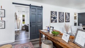 Una sala de estar moderna con una puerta corredera gris oscuro, una mesa de comedor de madera con un florero y un letrero de bienvenida enmarcados, y arte con motivos de hojas montado en la pared.